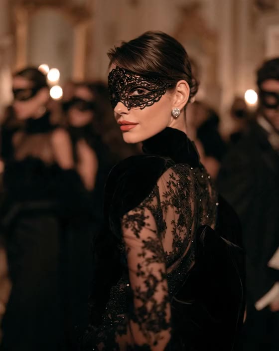 A woman in a black lace mask and sheer embroidered gown looks back over her shoulder in a candlelit ballroom