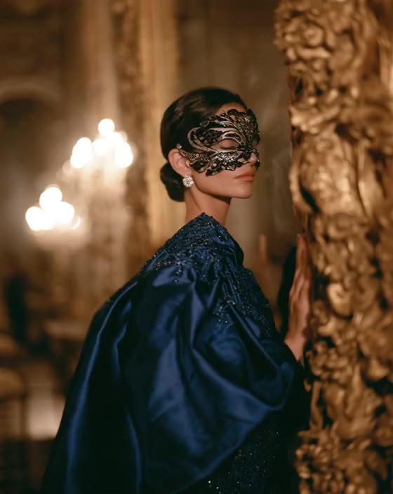 A woman in a midnight-blue satin gown with an open back walks past gilded mirrors
